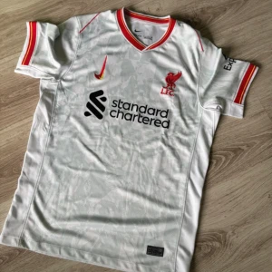 Liverpool FC vit bortatröja Nike - Liverpool FC:s officiella bortatröja från Nike. Tröjan är vit med subtilt mönster, röda och gula detaljer på krage och ärmslut. Broderad klubbmärke och Nike-logga på bröstet, samt sponsortryck. Tillverkad i lätt och ventilerande polyester.