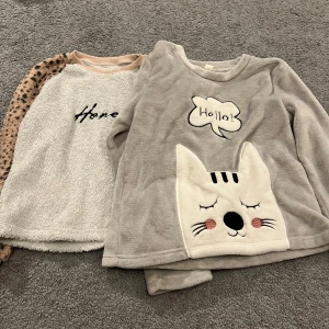 Två mysiga fleece-tröjor med print - Säljer två fluffiga fleece-tröjor. Ena är ljusgrå med ett sött kattansikte och texten 'Hello!' framtill, långärmad och supermjuk. Den andra är vit med texten 'Honey' och har beige leopardmönstrade ärmar. Perfekt för chill dagar hemma eller när du vill vara extra cozy.