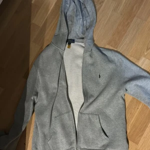 Grå hoodie från Polo Ralph Lauren XL - Säljer en grå hoodie från Polo Ralph Lauren i storlek S. Tröjan har huva, dragkedja och två fickor framtill. Klassisk Polo-logga broderad i marinblått på bröstet. Mjuk insida och skön passform, perfekt för chill dagar. Materialet är bomull och polyester.