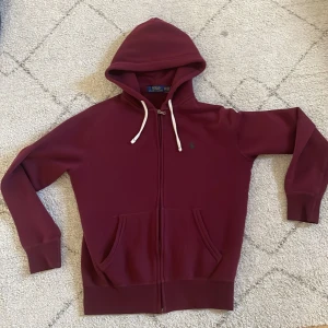 Vinröd hoodie från Polo Ralph Lauren - Snygg vinröd hoodie från Polo Ralph Lauren med dragkedja, huva med vita snören och två stora fickor framtill. Klassisk design med lång ärm och mjukt material som känns riktigt skönt. Perfekt för dig som gillar stilrena och bekväma plagg.