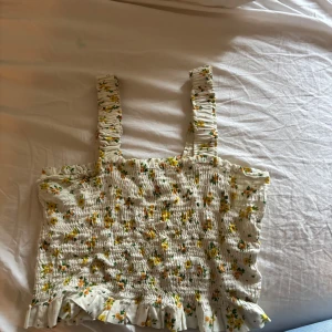 Blommig smockad linnetopp med volang - Supersöt linnetopp med smockad resår, breda axelband och volangkant nertill. Vit botten med små gula och orange blommor och gröna blad. Perfekt för soliga dagar! Den är fån veromoda och har endast testats på!💕