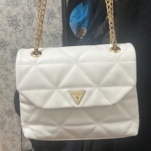 Vit quiltad axelväska från Guess - Supersnygg vit axelväska från Guess med quiltad design och guldfärgade kedjehandtag. Väskan har ett triangelformat Guess-emblem framtill och praktiskt innerfack med dragkedja. Perfekt storlek för det viktigaste och ger en clean vibe till din outfit. Dock står det nått på arabiska inne i väskan. Priset kan sänkas om nån är intresserad 