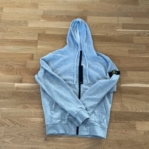 Grå hoodie från Stone Island - Snygg ljusgrå hoodie från Stone Island med klassisk logopatch på ärmen. Tröjan har huva med snörning, dragkedja framtill och två fickor. Mjuk bomullskvalitet och ribbade muddar vid ärmslut och nederkant. Perfekt för chill dagar och streetwear.