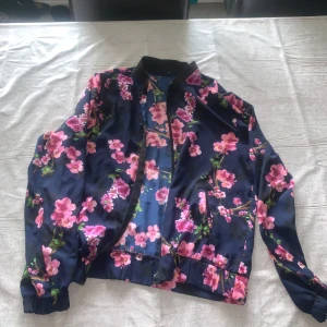 Blommig bomberjacka i mörkblått - Snygg bomberjacka med mörkblå bas och rosa blommor över hela jackan. Jackan har svarta muddar vid hals och ärmslut, samt dragkedja framtill. Perfekt för dig som vill sticka ut med ett färgglatt och trendigt plagg. Storlek L, men passar M