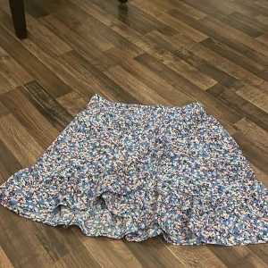 Blommig blå volangkjol - Supersöt kort kjol med blommigt mönster i blått, rosa, vitt och grönt. Kjolen har volangdetaljer och ett flowy, luftigt tyg som känns perfekt för varma dagar. Resår i halva midjan gör den enkel att dra på och skön att bära. Passar dig som gillar färg och mönster!