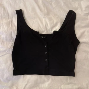 Svart croppad linnetopp med knappar - Snygg svart croppad linnetopp med knappar framtill och breda axelband. Toppen har en lätt ribbad struktur och en djupare urringning. Perfekt att styla med höga jeans eller kjol för en chill look.