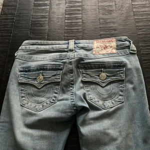 True Religion  - !!uppsydda i benen så de passar en person som är MAX 160!!Säljer ett par ljusblå True Religion Joey jeans med låg midja och bootcut. Klassiska bakfickor med lock och silverfärgade knappar med logga. Snygga kontrastsömmar och slitningseffekt ger en cool vintage vibe. Perfekta för dig som gillar streetstyle.