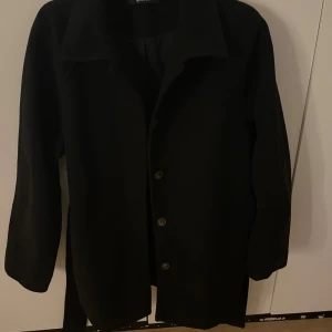 Svart kappa med knappar - Stilren svart kappa med klassisk krage och knäppning framtill. Jackan har långa ärmar och ett rakt snitt som ger en clean look. Perfekt för dig som gillar minimalistisk stil och vill ha något som funkar till många outfits.
