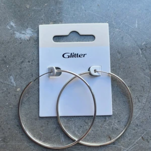 Stora silverfärgade hoops från Glitter - Säljer ett par stora, klassiska hoops i silverfärg från Glitter. Örhängena är runda och har en enkel, stilren design som ger en cool vibe till din look. Perfekt för dig som gillar statement-smycken och vill sticka ut lite extra.