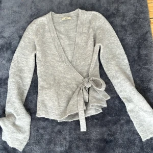 Grå omlottkofta från Gina Tricot - Supermjuk grå stickad omlottkofta från Gina Tricot med knyt i sidan och v-ringad hals. Långärmad och ribbade muddar för en chill och snygg look. Inga defekter och skicket är som nytt. 
