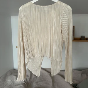 Plisserad offwhite blus med knyt - Superfin plisserad blus i offwhite med långa ärmar. Tunt och glansigt material med två knyten i ryggen. Aldrig använt med prislappen kvar!!