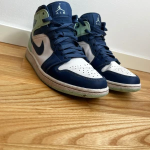 Nike Air Jordan 1 Mid blå/vit/grön - Nike Air Jordan 1 Mid i snygg färgkombination av marinblått, vitt och mintgrönt. Klassisk high-top siluett med snörning, rund tå och vit sula. Ovandel i skinn och syntet, ikoniska Jordan Wings-loggan på sidan och Jumpman på plösen. Perfekta för dig som gillar streetstyle. Väldigt bra skick förutom den lilla skadan på vänster sko. 