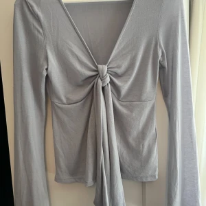 Ljusblå långärmad topp Gina Tricot - Supersnygg ljusblå (åt det lila hållet) långärmad topp från Gina Tricot med djup v-ringning och knyt framtill. Toppens material är mjukt och stretchigt, perfekt för en trendig look. Passformen är normal och toppen har en stilren, enkel design som är lätt att matcha.