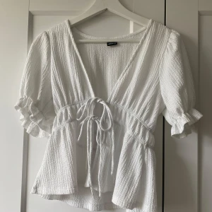 Vit blus med knyt från Gina Tricot - Supersöt vit blus från Gina Tricot i storlek XS. Blusen har puffiga korta ärmar med volangkant, v-ringning och snygg knytning i midjan. Materialet är strukturerat och känns luftigt, perfekt för vår och sommar. Passformen är normal och blusen har en feminin siluett.