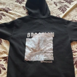 Svart hoodie med Brooklyn-tryck - Svart hoodie med stor huva och snörning, samt känguruficka framtill. Baksidan har ett stort tryck med texten 'BROOKLYN BRIDGE' och en bild av Brooklyn Bridge. Tröjan är i mjuk bomullsmix och har ribbade muddar vid ärmslut och nederkant. Black sweatshirt for men and woman. With a text and a picture on the back “brocklyn bringe” with a photo of Brooklyn. It’s a size M. Very warm and great for fall and winter❄️Gives a comfy look and goes with everything.