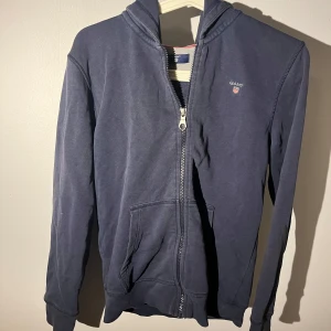 Mörkblå hoodie från GANT med dragkedja - Säljer en mörkblå hoodie från GANT med huva och praktisk dragkedja framtill. Skicket är sådär då den är lite uttvättad men inga hål eller skador så. Storleken är xs och sitter lite tajtare. 