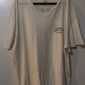 Vit/Beige Lee t-shirt med tryck - Snygg vit/beige t-shirt från Lee med coolt Kansas Outlaws-tryck och örn på ryggen. Rund hals och korta ärmar, tillverkad i mjuk bomull. Perfekt för dig som gillar streetwear och vill ha en chill vibe. Tryck även fram på bröstet.