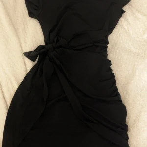 Nyköpt svart kortklänning med knyt i midjan - Snygg svart kortklänning med knyt i midjan som ger en cool och figurnära siluett. Klänningen har korta ärmar och är perfekt för dig som gillar enkel men stilren design. Passar dig som vill ha en klassisk look med en twist.