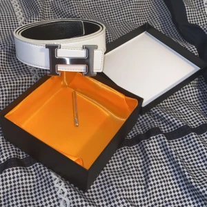  vitt skärp med H-spänne - Snyggt vitt bälte från med ikoniskt H-spänne i silverfärg. Bältet är gjort i skinn och har en stilren, klassisk design med svarta kanter. Perfekt accessoar för att lyfta din outfit med en lyxig touch.