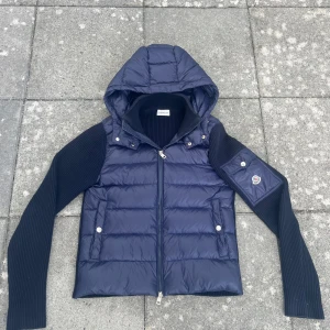 Mörkblå Moncler pufferjacka med huva - Snygg mörkblå pufferjacka (cardigan) från Moncler med stickade ärmar och avtagbar huva. Jackan har dragkedja framtill, två fickor med tryckknappar och Moncler-logga på ärmen. Perfekt för kyliga dagar och riktigt stilren look. nyskick lappar följer med etc. Passar båda man och kvinna