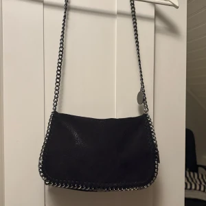 Svart axelväska med kedjerem - Snygg svart axelväska med kedjerem och coola metallkedjor längs kanterna. Väskan har en mjuk, mockaliknande yta och en stilren, enkel form som passar till många outfits. Perfekt för dig som gillar en edgy och trendig look. Köpt här på Plick❣️