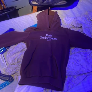Brun Peak Performance hoodie - Säljer en brun hoodie från Peak Performance med vit broderad logga på bröstet. Klassisk modell med huva och långa ärmar, perfekt för chill dagar. Mjuk insida och ribbade muddar för extra komfort.