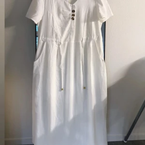 Vit långklänning med knappar - Snygg vit långklänning från Indiska med korta ärmar, v-ringning och dekorativa knappar framtill. Klänningen har dragsko i midjan med guldfärgade detaljer och praktiska fickor på sidorna. Perfekt för sommarens alla tillfällen.