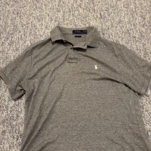 Grå pikétröja från Polo Ralph Lauren - Snygg grå pikétröja från Polo Ralph Lauren med klassisk krage och två knappar framtill. Tröjan har korta ärmar och den ikoniska broderade loggan på bröstet. Custom fit-modell i mjuk bomull som är skön att ha på sig hela dagen.