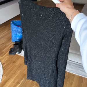 Svart glittrig omlottklänning - Snygg svart kortklänning med glittrigt tyg och omlottmodell. Klänningen har djup urringning i ryggen med knytband och långa ärmar. Perfekt för dig som vill sticka ut med lite extra glitter. Aldrig använd