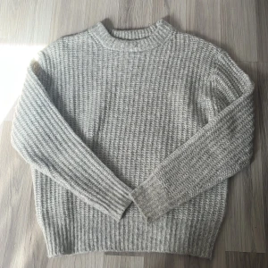 Grå stickad tröja med ribbad struktur - Säljer en grå stickad tröja med ribbad struktur. Tröjan har långa ärmar och är perfekt för kyliga dagar. Den är mjuk och har en klassisk, enkel design som funkar till allt. Passformen är normal och materialet känns riktigt mysigt. Endast använd en gång.