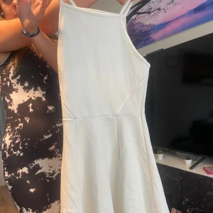 Vit kortklänning med smala axelband - Säljer en fräsch vit kortklänning med smala axelband och lätt utställd kjol. Klänningen har ett diskret mönster i tyget och är perfekt för varma dagar. Enkel och stilren design som är lätt att matcha med accessoarer.