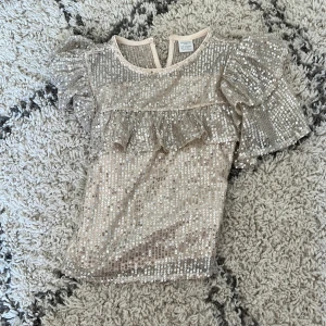 Glittrig volangblus från Lindex Kids - Superfin glittrig blus från Lindex Kids i ljusbeige med massor av paljetter❤️. Blusen har korta volangärmar ❤️tröja är i bra skick och sälja för att den är för liten ❤️