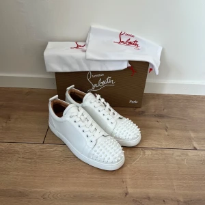 Christian Louboutin vita sneakers med nitar - Säljer ett par sjukt snygga vita sneakers från Christian Louboutin med coola vita nitar på tån. Skorna har snörning, rund tå och är tillverkade i lyxigt skinn. Perfekt för dig som vill sticka ut med en exklusiv vibe och edgy detaljer.