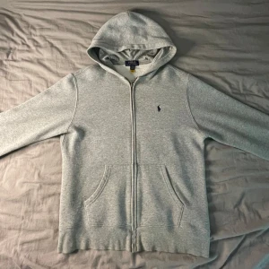 Grå Zip-Up Hoodie från Polo Ralph Lauren - Säljer en grå zip-up hoodie från Polo Ralph Lauren. Tröjan är sparsamt använd, inga slitage märken men som man kan se på bilderna, syns de att den har blivit tvättad ett flertal gånger, längst ner på tröjan.  Storlek 160 (L/G 14-16)  Mått 📏📐 - Bröstvidd 48cm  - Bottenvidd 44cm - Armlängd 43cm - Plagglängd 57cm  Hör gärna av er vid frågor  ~ LoopSupply ~