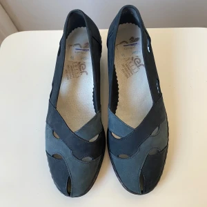 Svarta ballerinaskor från Rieker - Snygga ballerinaskor från Rieker i mörk mocka med coola utskurna detaljer och rund tå. Skorna har en bekväm passform, perfekta för dig som gillar stilren design med en twist.