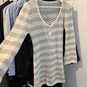 Vit och beige stickad kortklänning - Säljer en söt stickad kortklänning i vit och beige med långärmade armar och v-ringad hals. Klänningen har ett genomgående hålmönster och är ribbad vid halsen. Strandklänning 