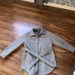 Grå kappa men knappar  - Snygg grå overshirt med bälte i midjan och klassisk krage. Jackan har knappar framtill och långa ärmar, perfekt för lager-på-lager-stil. Materialet känns mjukt och passar både till jeans och kjol.