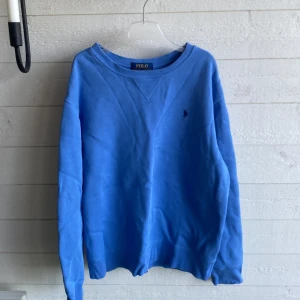 Blå sweatshirt från Polo Ralph Lauren - Blå sweatshirt från Polo Ralph Lauren med klassisk rund hals och broderad logga på bröstet. Tröjan har långa ärmar och ribbade muddar vid ärmslut och nederkant. Perfekt för chill dagar och enkel att matcha med jeans eller joggers.