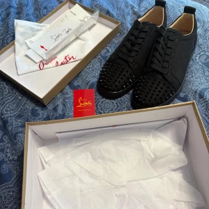 Christian Louboutin svarta sneakers med nitar - Säljer ett par svarta sneakers från Christian Louboutin med coola svarta nitar på tån och klassisk röd sula med logga. Skorna har snörning och är tillverkade i mocka med läderdetaljer på insidan. Perfekt för dig som vill sticka ut med exklusiv stil.