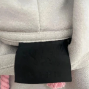 Grå hoodie med rosa tryck från Syna world  - Säljer en grå hoodie från SANTA med fett rosa graffiti-tryck på huvan och grova rosa snören. Tröjan har en klassisk känguruficka och mjukt material som känns skönt mot huden. Perfekt för chill dagar och streetwear vibes. Pris kan snackas vid snabba affärer 