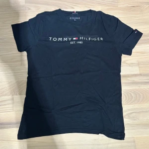  Tommy Hilfiger t-shirt svart - Svart t-shirt från Tommy Hilfiger med klassisk logga och texten 'EST. 1985' tryckt på bröstet. Gjord i mjuk ekologisk bomull, rund hals och korta ärmar. Snygg och enkel design med liten Hilfiger-detalj på ärmen. Perfekt för dig som gillar stilrena plagg.