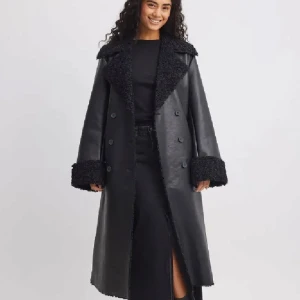 Svart lång pälsjacka i skinnlook - Lång svart fuskpälsfodrad kappa – Helt ny Snygg och stilren lång kappa i svart med mjukt, värmande fuskpälsfoder och detaljer vid krage och ärmslut. Dubbelknäppning framtill och en rak, elegant passform som passar perfekt till både vardag och fest.  Anledning till försäljning: För stor och hann inte returnera i tid.  Perfekt för dig som vill ha en trendig och varm kappa till höst och vinter!