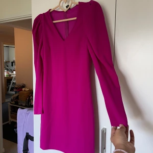 Fuchsia kortklänning med puffärm - Säljer en snygg fuchsiafärgad kortklänning med v-ringning och långa puffärmar. Klänningen har en stilren och figurnära passform, perfekt för dig som gillar färg och vill sticka ut. Materialet känns lätt och mjukt mot huden.Helt ny