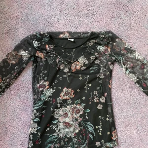 Blommig meshblus med volang från Lindex - Supersöt svart meshblus från Lindex med blommigt mönster i rosa, grönt och vitt. Blusen har långa transparenta ärmar och volangdetalj längs ringningen. Perfekt för dig som gillar romantisk och trendig stil. Den är i storlek 146-152 cm och är i väldigt bra skick.