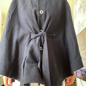 Svart kappa med knytband PONCHO - Stilren svart kappa med knytband i midjan och snygga guldiga knappdetaljer. Jackan har en rak passform, stora fickor och är tillverkad i ett mjukt ullmaterial. Perfekt för dig som vill ha en klassisk look med en twist.