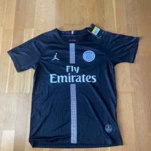 PSG x Jordan svart matchtröja 2018/2019 - Svart PSG fotbollströja i samarbete med Jordan, med vit logga och emblem på bröstet samt Fly Emirates-tryck. Tröjan har korta ärmar och en smal vertikal rand i mitten.