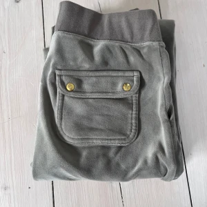 Grå-gröna juicybyxor - Snygga grå-gröna juicy byxor  med bred resår och snörning i midjan, guldfärgade detaljer och praktiska fickor både fram och bak. Mjukt material som känns skönt mot huden och broderad logga framtill. Perfekta för chill eller streetwear. Bra skick. inga defekter