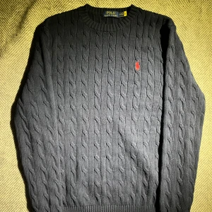 Mörkblå kabelstickad tröja från Polo Ralph Lauren - Säljer en stilren mörkblå kabelstickad tröja från Polo Ralph Lauren med det klassiska röda logotypbroderiet på bröstet. Tröjan har långa ärmar och en rund halsringning. Perfekt för en elegant och avslappnad look.