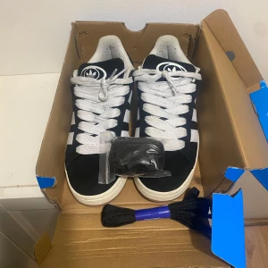 Svarta Adidas Campus sneakers - Svarta Adidas Campus sneakers med klassiska vita ränder och stor vit logga på plösen. Skorna har breda vita snören och kommer med extra svarta skosnören. Ovandel i mocka och sula i vitt gummi. Perfekt för dig som gillar retrostil och streetwear. Söker byte!