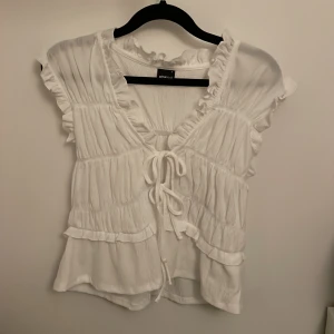 Vit blus med volanger från Gina Tricot - Supersöt vit blus från Gina Tricot med volangdetaljer och knytning framtill. Blusen har v-ringning, ärmlös design och ett lätt, luftigt tyg. Endast använd en gång och säljer pågrund av att den inte kommer till användning💗